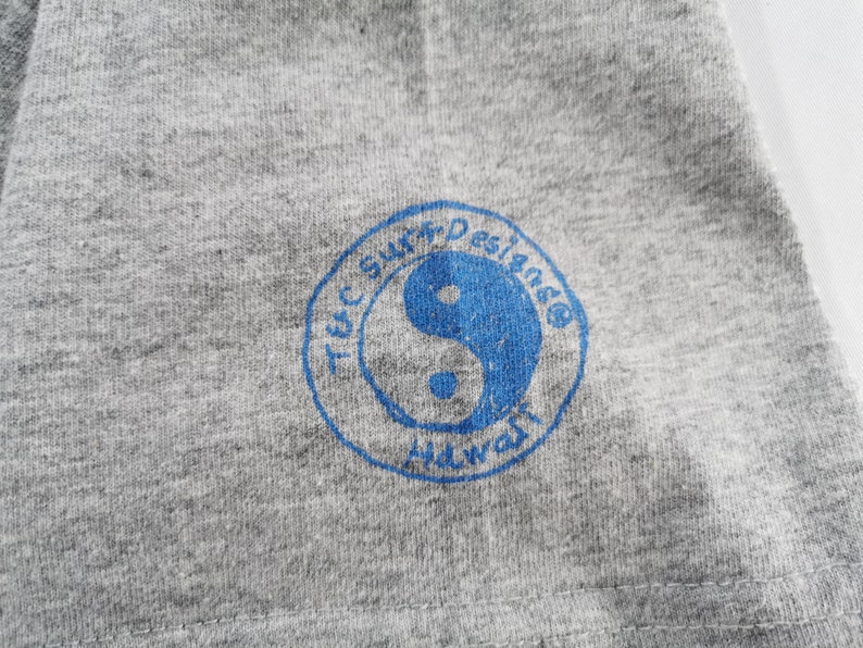 May include: Close-up of a gray t-shirt with a blue circular logo. The logo features a yin-yang symbol and the text "T&C Surf Designs Hawaii". The fabric has a heathered texture.