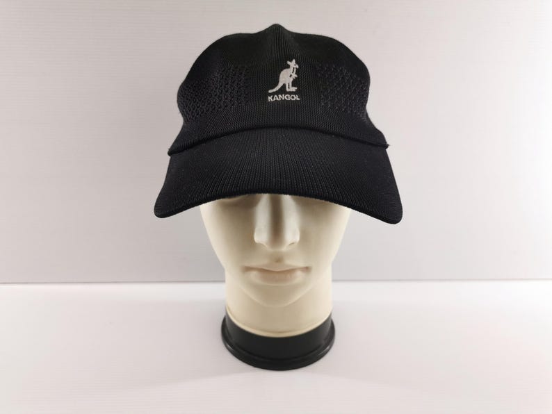 Puede incluir: Gorra Kangol negra con visera curva, exhibida en una cabeza de maniqu&iacute;. La gorra presenta el logotipo de Kangol en blanco. La gorra est&aacute; hecha de un material texturizado y transpirable.