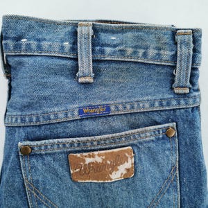 May include: Close-up of the back of blue denim Wrangler jeans. The jeans have a classic five-pocket design with a brown leather patch and the Wrangler logo. The jeans have belt loops and a button closure.