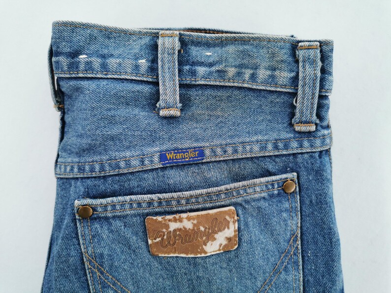 May include: Close-up of the back of blue denim Wrangler jeans. The jeans have a classic five-pocket design with a brown leather patch and the Wrangler logo. The jeans have belt loops and a button closure.
