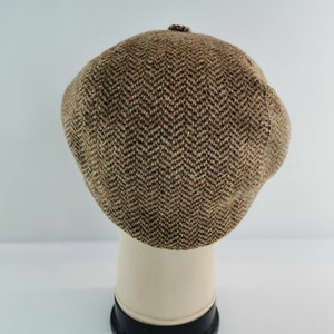 Op de afbeelding: Een bruin en tan visgraatpatroon newsboy pet. De pet wordt getoond op een witte mannequin hoofd met een zwarte basis. De pet heeft een kleine knoop aan de bovenkant. De pet is gemaakt van een tweed-achtig materiaal.