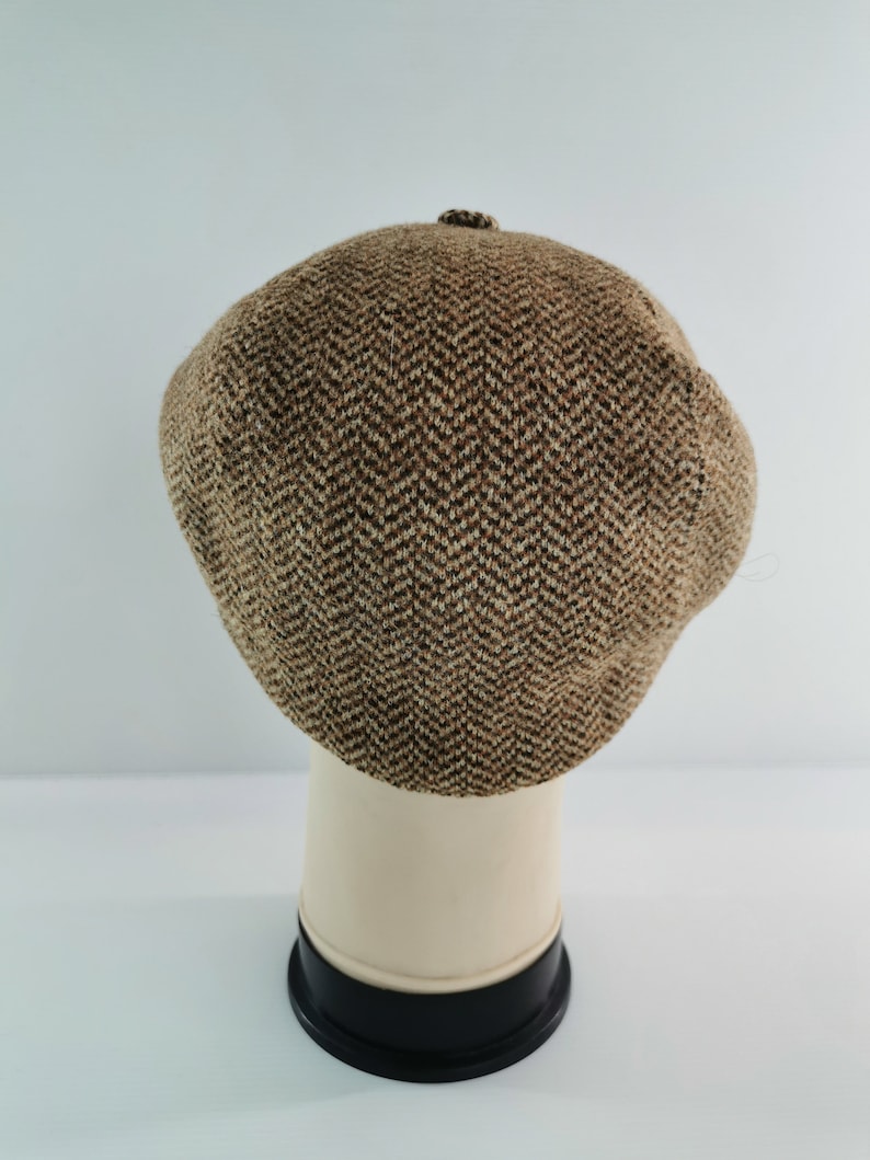 Op de afbeelding: Een bruin en tan visgraatpatroon newsboy pet. De pet wordt getoond op een witte mannequin hoofd met een zwarte basis. De pet heeft een kleine knoop aan de bovenkant. De pet is gemaakt van een tweed-achtig materiaal.