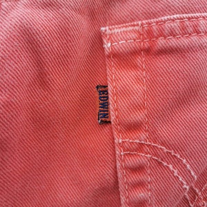 Op de afbeelding: Close-up van koraalkleurige denimstof, met details van de zak en stiksels. Een klein, donkerblauw label met de merknaam "EDWIN" is zichtbaar. De stof heeft een getextureerde uitstraling.