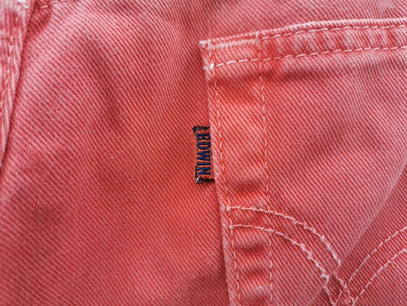 Op de afbeelding: Close-up van koraalkleurige denimstof, met details van de zak en stiksels. Een klein, donkerblauw label met de merknaam "EDWIN" is zichtbaar. De stof heeft een getextureerde uitstraling.