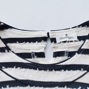 Puede incluir: Primer plano de un top color crema con rayas horizontales azul marino. El top tiene un cuello redondo con una abertura de ojo de cerradura en la espalda. El nombre de la marca "courr&egrave;ges" est&aacute; impreso en una peque&ntilde;a etiqueta.