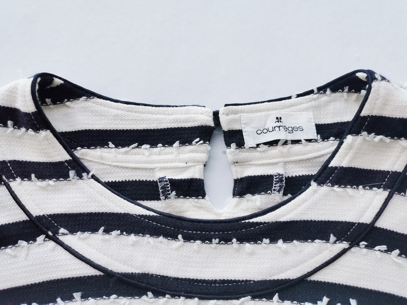 Puede incluir: Primer plano de un top color crema con rayas horizontales azul marino. El top tiene un cuello redondo con una abertura de ojo de cerradura en la espalda. El nombre de la marca "courr&egrave;ges" est&aacute; impreso en una peque&ntilde;a etiqueta.