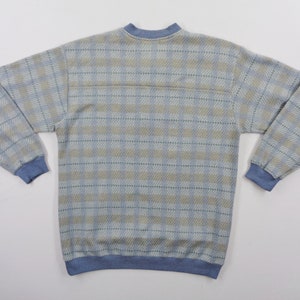 Puede incluir: Sudadera con estampado de cuadros en azul claro y beige con pu&ntilde;os, cuello y dobladillo azules. La parte superior de manga larga tiene cuello redondo y un aspecto suave y texturizado. La parte posterior de la camisa es visible.