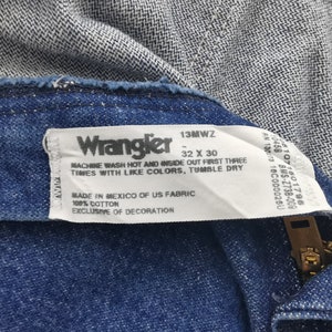 K&ouml;nnte beinhalten: Nahaufnahme einer blauen Wrangler-Jeans, die das Etikett mit dem Markennamen, der Gr&ouml;&szlig;e 32 x 30 und Pflegehinweisen zeigt. Die Jeans besteht aus 100 % Baumwollstoff.
