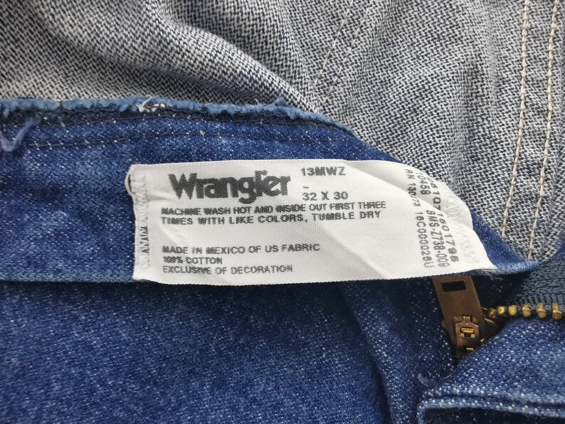 K&ouml;nnte beinhalten: Nahaufnahme einer blauen Wrangler-Jeans, die das Etikett mit dem Markennamen, der Gr&ouml;&szlig;e 32 x 30 und Pflegehinweisen zeigt. Die Jeans besteht aus 100 % Baumwollstoff.