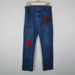 May include: Blue denim jeans featuring a high-waisted cut, a button and zipper fly closure, and belt loops. The garment includes a dark brown square patch on the upper left thigh and a smaller red square patch on the lower right leg.