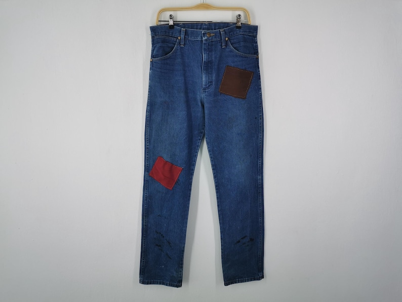 May include: Blue denim jeans featuring a high-waisted cut, a button and zipper fly closure, and belt loops. The garment includes a dark brown square patch on the upper left thigh and a smaller red square patch on the lower right leg.