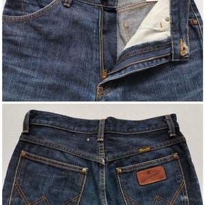 May include: Dark wash denim Wrangler jeans. The jeans have a classic five-pocket design with a zip fly and button closure. The back pockets feature the signature Wrangler stitching and a leather patch with the Wrangler logo.