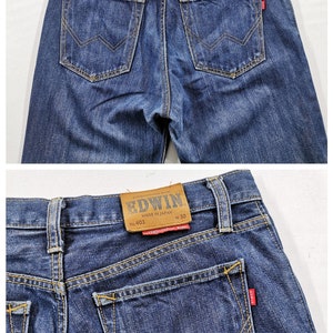 May include: Close-up of the back of blue denim jeans. The jeans have two back pockets with a distinctive W-shaped stitch design. A brown leather patch with the brand name "EDWIN" is visible at the waistband.