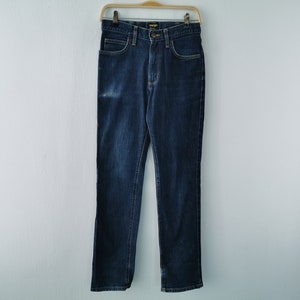 May include: Dark blue denim jeans hanging on a wooden hanger. The jeans have a classic five-pocket design, a button and zipper closure, and the Wrangler brand label. The jeans are a straight-leg style.