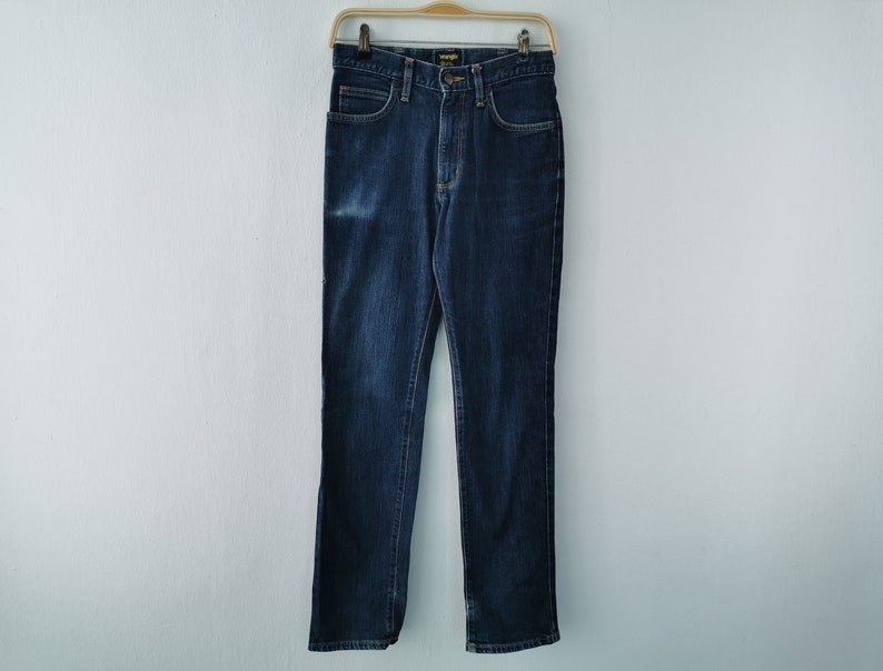 May include: Dark blue denim jeans hanging on a wooden hanger. The jeans have a classic five-pocket design, a button and zipper closure, and the Wrangler brand label. The jeans are a straight-leg style.
