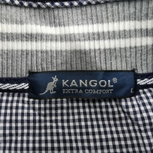 May include: Close-up of a gray and white striped shirt with a navy blue label that reads "KANGOL EXTRA COMFORT" and a kangaroo logo. The shirt has a checkered pattern on the bottom.