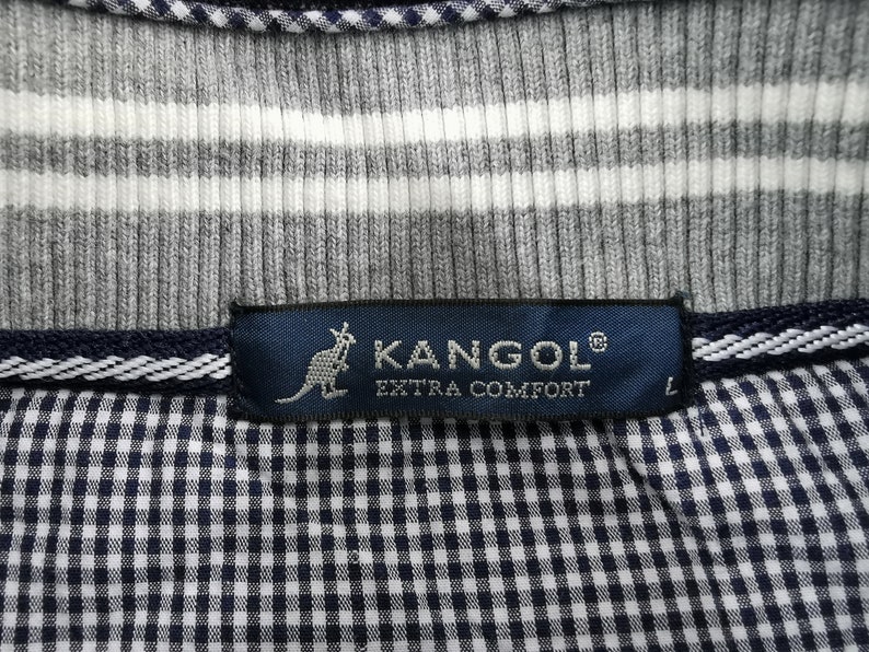 May include: Close-up of a gray and white striped shirt with a navy blue label that reads "KANGOL EXTRA COMFORT" and a kangaroo logo. The shirt has a checkered pattern on the bottom.