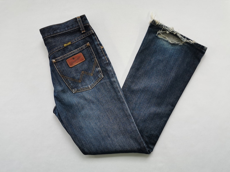 May include: Dark wash denim jeans with a classic five-pocket design. Features a brown leather patch with the Wrangler logo on the back pocket. The jeans have a distressed hem, adding a vintage look. The jeans are folded neatly.