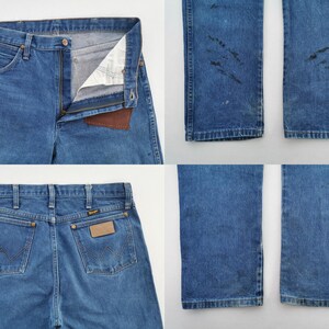 May include: Blue denim jeans featuring a front zipper closure, a brown leather patch on the interior waistband, and two back pockets with a stitched W design. The legs show dark distressed markings and wear patterns on the lower sections.