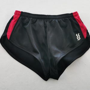 May include: Black athletic shorts featuring a high-cut leg design with vibrant red side panels and an elastic waistband. A small white graphic logo consisting of two vertical wavy lines is positioned on the lower right side of the garment.