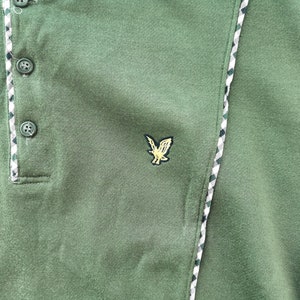 May include: Green polo shirt with a three-button placket and an embroidered eagle logo. The shirt has a textured fabric and a decorative trim along the placket and side seam. The trim features a pattern of white, green, and black.