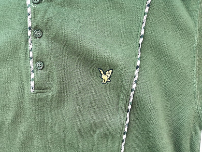 May include: Green polo shirt with a three-button placket and an embroidered eagle logo. The shirt has a textured fabric and a decorative trim along the placket and side seam. The trim features a pattern of white, green, and black.