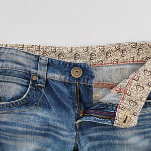 May include: Close-up of blue denim jeans with a button closure and zipper. The jeans have a decorative patterned lining visible at the waistband and pocket. The denim has a faded, worn look.