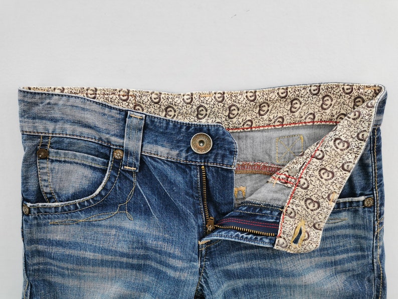 May include: Close-up of blue denim jeans with a button closure and zipper. The jeans have a decorative patterned lining visible at the waistband and pocket. The denim has a faded, worn look.