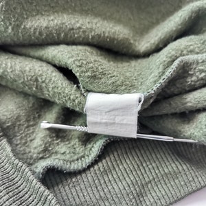 May include: Close-up of olive green sweatshirt material. The fabric is folded, showing a tag and a silver pin. The texture of the material is visible, with a ribbed knit cuff and a soft, fleecy interior.