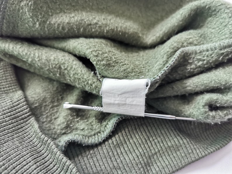 May include: Close-up of olive green sweatshirt material. The fabric is folded, showing a tag and a silver pin. The texture of the material is visible, with a ribbed knit cuff and a soft, fleecy interior.