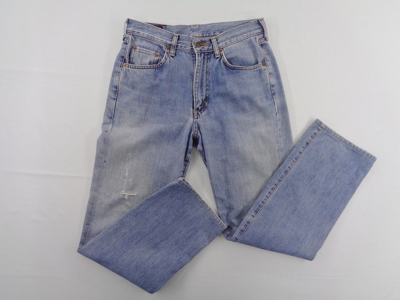 May include: Light blue denim jeans with a classic five-pocket design. The jeans have a faded wash and a distressed detail on the front. The jeans are folded and laid flat on a white surface.