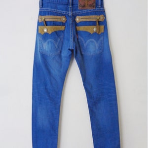 K&ouml;nnte beinhalten: Blaue Jeans mit braunen Wildleder-&auml;hnlichen Akzenten auf den Ges&auml;&szlig;taschen und Rei&szlig;verschl&uuml;ssen. Die Jeans haben einen Lederaufn&auml;her am Bund und ein rotes Etikett auf der Ges&auml;&szlig;tasche. Die Jeans h&auml;ngen an einem Kleiderb&uuml;gel.