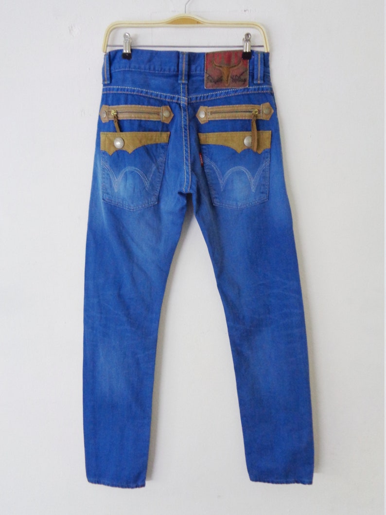 K&ouml;nnte beinhalten: Blaue Jeans mit braunen Wildleder-&auml;hnlichen Akzenten auf den Ges&auml;&szlig;taschen und Rei&szlig;verschl&uuml;ssen. Die Jeans haben einen Lederaufn&auml;her am Bund und ein rotes Etikett auf der Ges&auml;&szlig;tasche. Die Jeans h&auml;ngen an einem Kleiderb&uuml;gel.