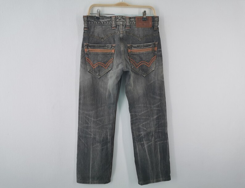 May include: Gray denim jeans with orange stitching accents on the back pockets and seams. The jeans are hanging on a wooden hanger against a white background. The jeans have a faded, worn look.