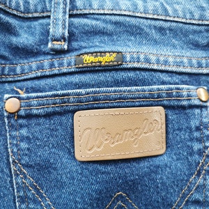May include: Close-up of blue denim jeans with the Wrangler logo on the back pocket. The jeans have copper-colored rivets and stitching. The Wrangler label is black with gold lettering.