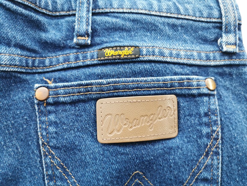 May include: Close-up of blue denim jeans with the Wrangler logo on the back pocket. The jeans have copper-colored rivets and stitching. The Wrangler label is black with gold lettering.