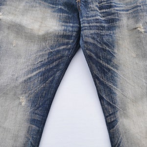 Pu&ograve; includere: Primo piano di jeans di denim blu strappati. I jeans presentano aree sbiadite e sottili motivi di usura, con alcuni piccoli strappi. Il tessuto mostra un mix di tonalit&agrave; blu chiare e scure, creando un look vintage.