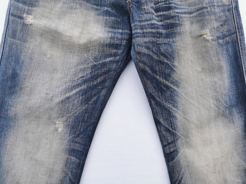 Pu&ograve; includere: Primo piano di jeans di denim blu strappati. I jeans presentano aree sbiadite e sottili motivi di usura, con alcuni piccoli strappi. Il tessuto mostra un mix di tonalit&agrave; blu chiare e scure, creando un look vintage.