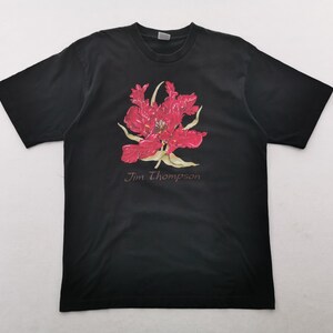 May include: Black t-shirt featuring a large, vibrant red flower graphic with yellow accents. The text "Jim Thompson" is printed below the floral design. The shirt is made of a soft material.