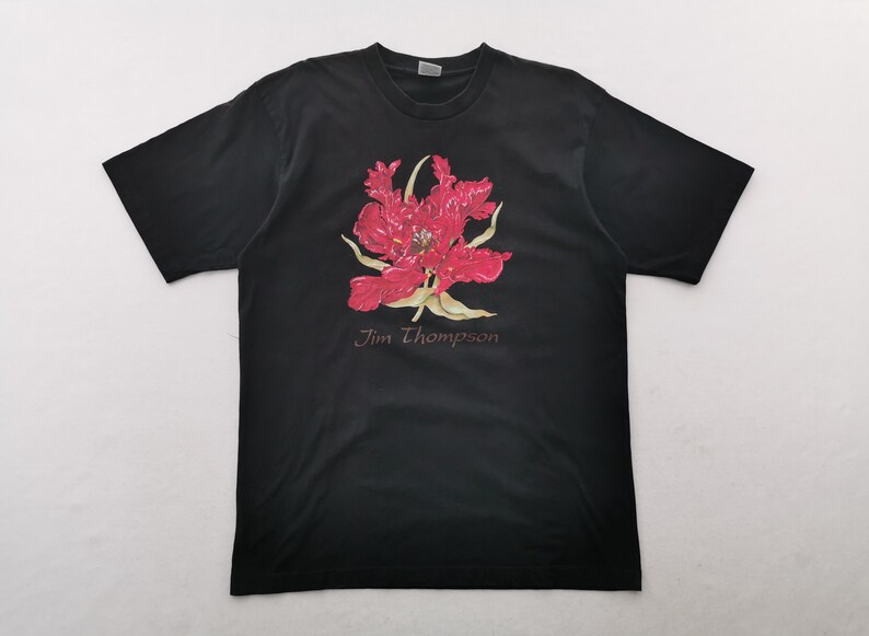 May include: Black t-shirt featuring a large, vibrant red flower graphic with yellow accents. The text "Jim Thompson" is printed below the floral design. The shirt is made of a soft material.