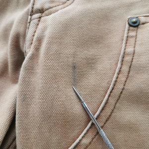 May include: Close-up of tan-colored fabric, possibly canvas or a similar material, showing detailed stitching and a metal button. A small, dark stain is visible on the fabric's surface. A silver tool is pointing at the fabric.