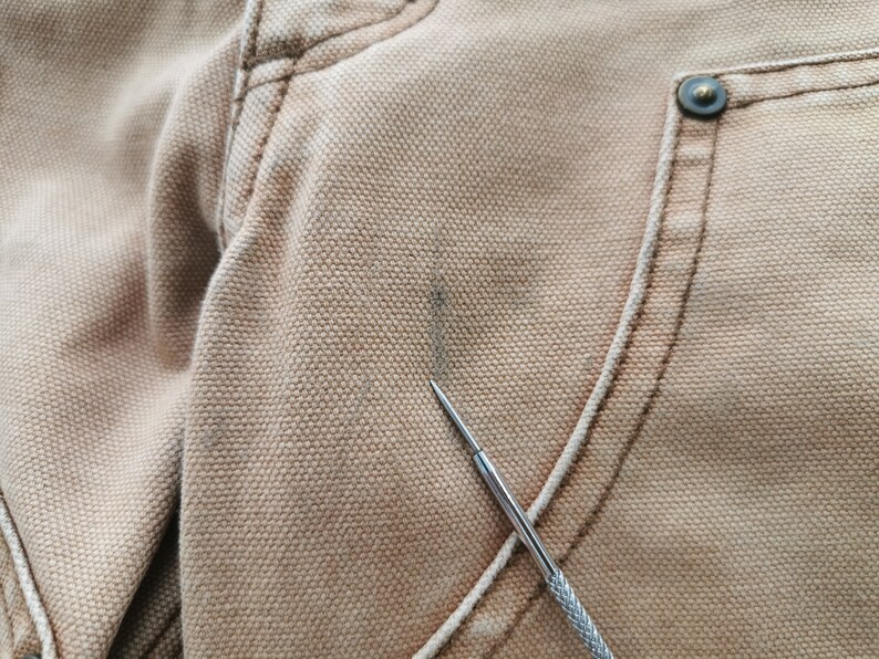 May include: Close-up of tan-colored fabric, possibly canvas or a similar material, showing detailed stitching and a metal button. A small, dark stain is visible on the fabric's surface. A silver tool is pointing at the fabric.