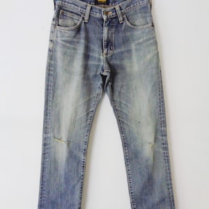 Pu&ograve; includere: Jeans in denim blu sbiadito con dettagli strappati, tra cui strappi sulle ginocchia. I jeans sono appesi a una gruccia su sfondo bianco. Il logo Wrangler &egrave; visibile sulla tasca posteriore.