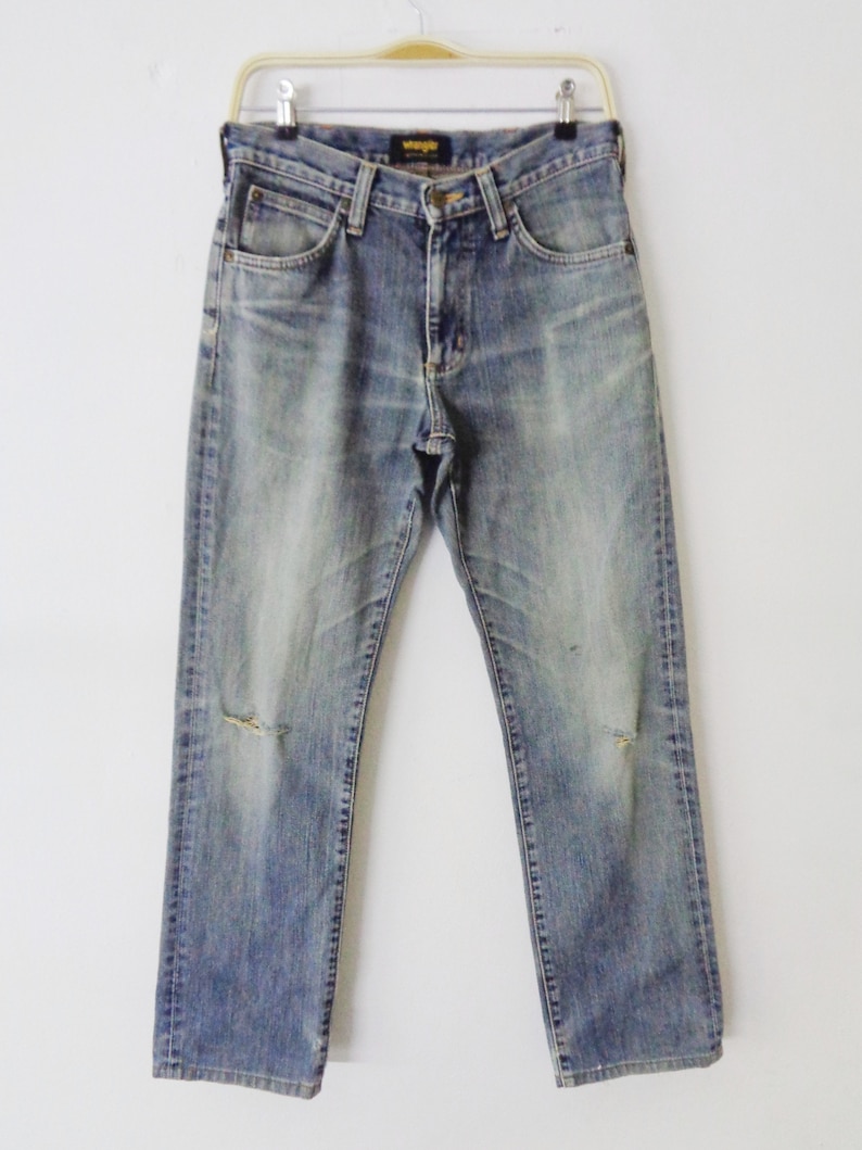 Pu&ograve; includere: Jeans in denim blu sbiadito con dettagli strappati, tra cui strappi sulle ginocchia. I jeans sono appesi a una gruccia su sfondo bianco. Il logo Wrangler &egrave; visibile sulla tasca posteriore.