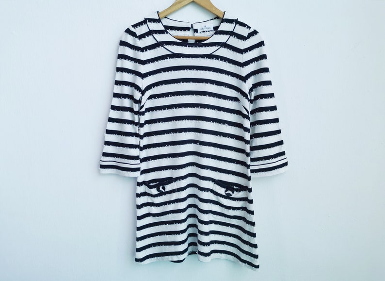 Puede incluir: Un vestido a rayas blancas y negras con mangas tres cuartos cuelga de una percha de madera. El vestido tiene un cuello redondo y dos bolsillos delanteros. Las rayas son horizontales y est&aacute;n espaciadas uniformemente.