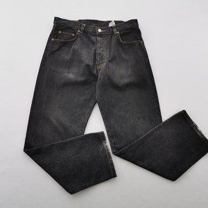 Puede incluir: Vaqueros de mezclilla gris oscuro con bragueta de botones, diseño de cinco bolsillos y pernera recta. Los vaqueros tienen un aspecto descolorido y están colocados planos. Las costuras son de color beige contrastante.