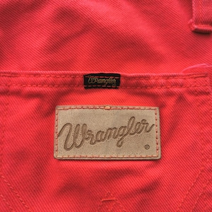 May include: Close-up of bright red denim pants with a back pocket and a Wrangler brand patch. The Wrangler logo is also on a small black tag above the pocket. The stitching is visible.