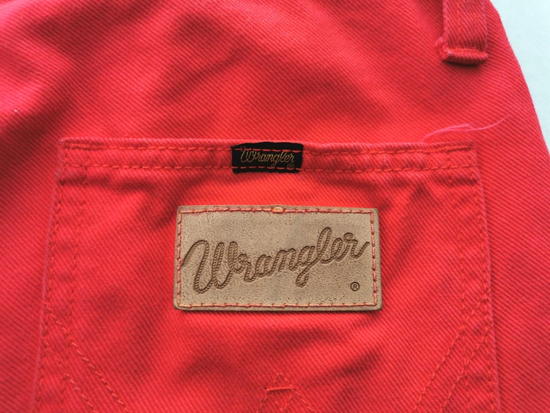 May include: Close-up of bright red denim pants with a back pocket and a Wrangler brand patch. The Wrangler logo is also on a small black tag above the pocket. The stitching is visible.