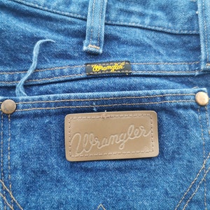 K&ouml;nnte beinhalten: Nahaufnahme der R&uuml;ckseite einer blauen Wrangler-Jeans. Die Jeans hat einen braunen Lederaufn&auml;her mit dem Wrangler-Logo, Kupfernieten und ein kleines schwarzes Etikett mit dem Wrangler-Logo. Die N&auml;hte sind in einem kontrastierenden Beigeton gehalten.