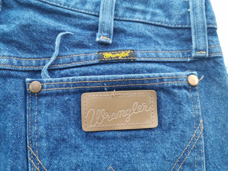 K&ouml;nnte beinhalten: Nahaufnahme der R&uuml;ckseite einer blauen Wrangler-Jeans. Die Jeans hat einen braunen Lederaufn&auml;her mit dem Wrangler-Logo, Kupfernieten und ein kleines schwarzes Etikett mit dem Wrangler-Logo. Die N&auml;hte sind in einem kontrastierenden Beigeton gehalten.